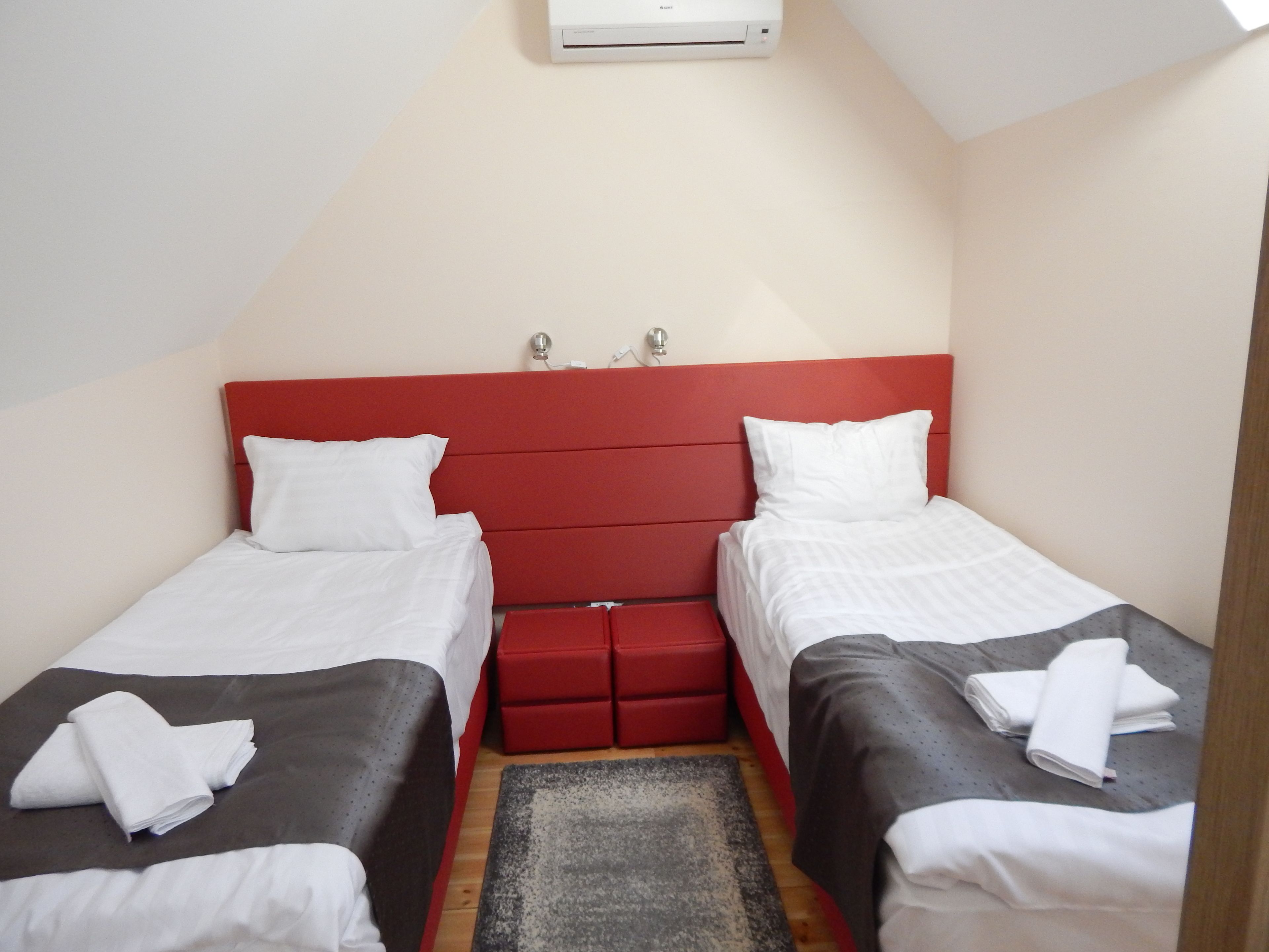 Une petite chambre d’hôtel, de taille modeste, avec deux lits simples, une tête de lit rouge et un petit banc rouge placé entre les lits.