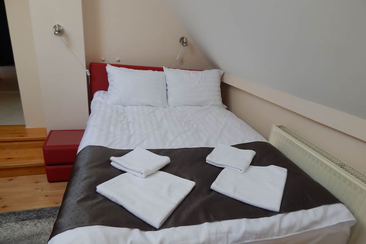 A neatly made bed with white linens and a brown accent, a red bedside table, and a white radiator.