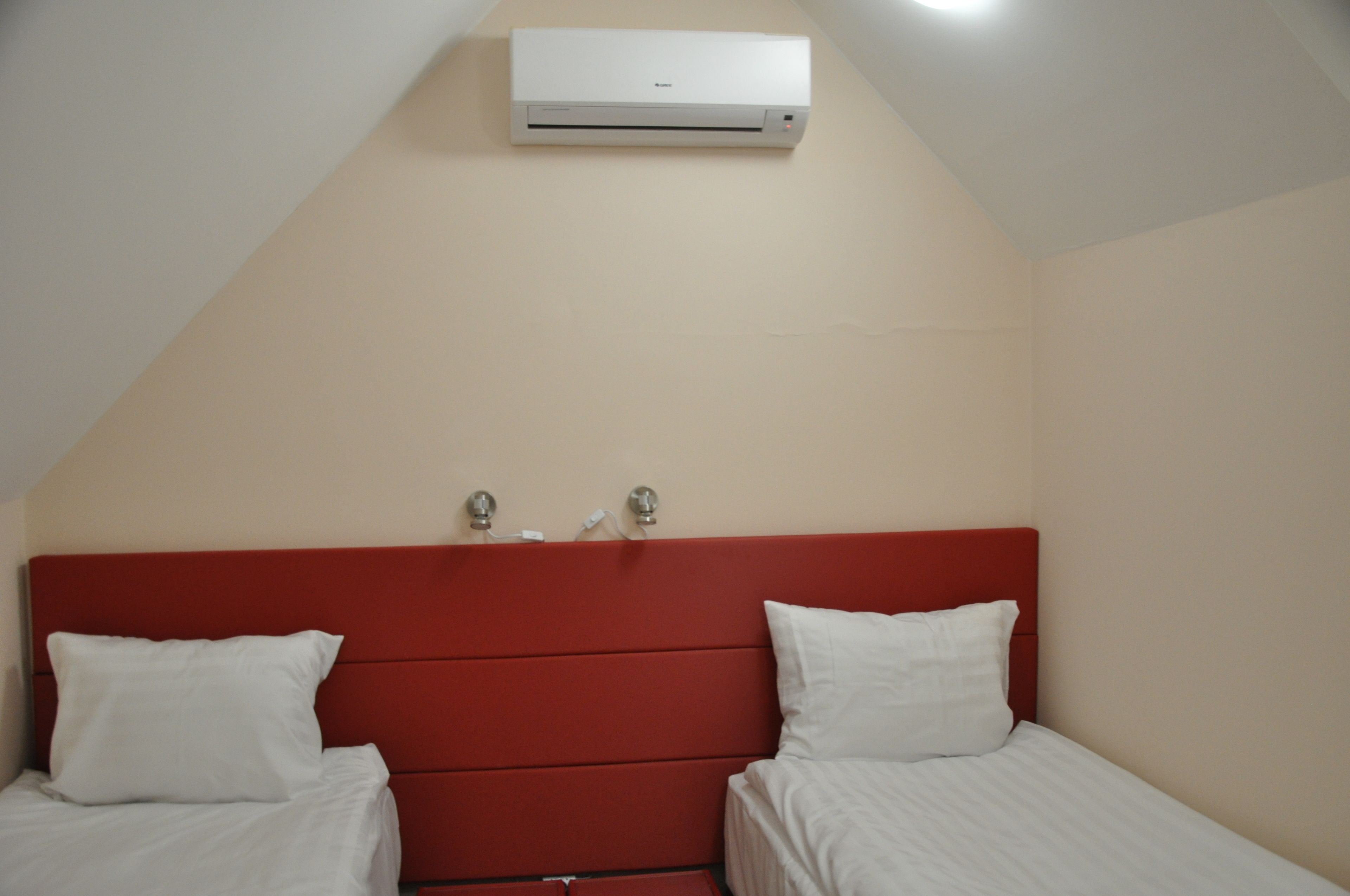 Een slaapkamer met een rode hoofdbord, twee eenpersoonsbedden, witte beddengoed en een airconditioningunit aan de muur.