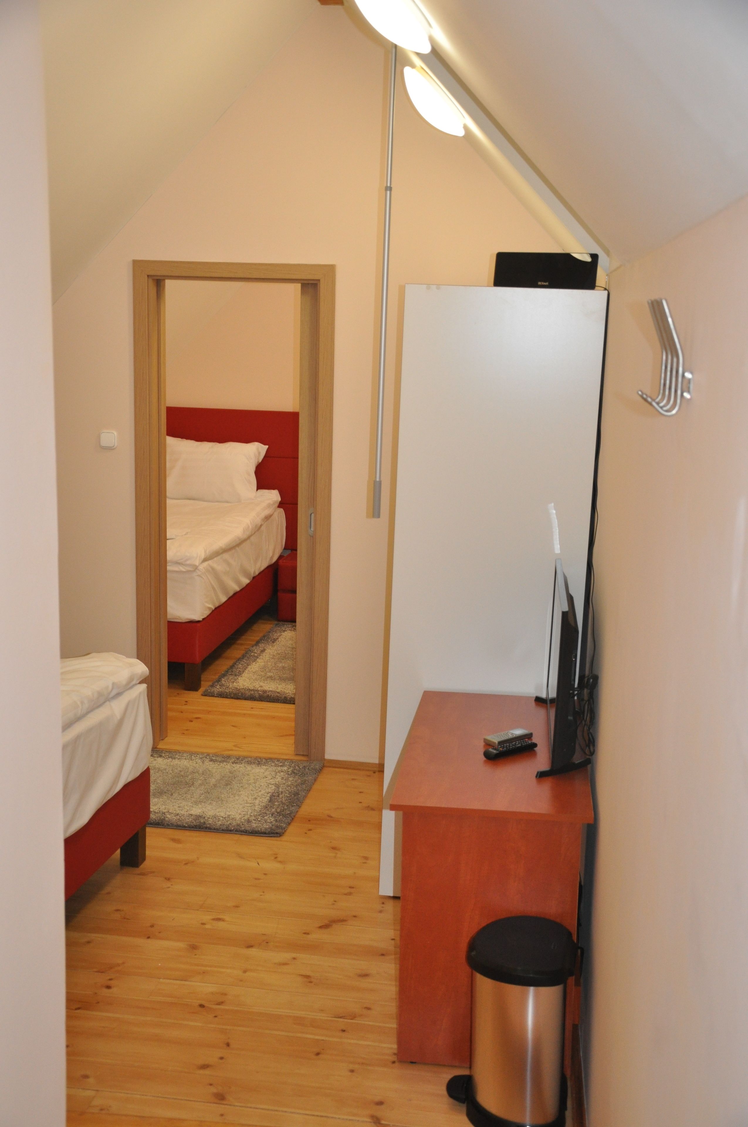 Una habitación pequeña y funcional con suelo de madera, una cama con ropa blanca, un escritorio con una computadora y un cubo de basura.