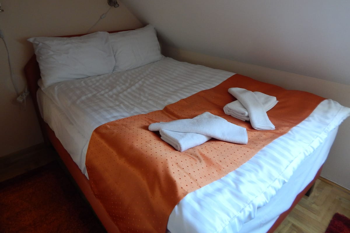 A neatly made bed with white linens, an orange accent throw, and folded towels on the bed.