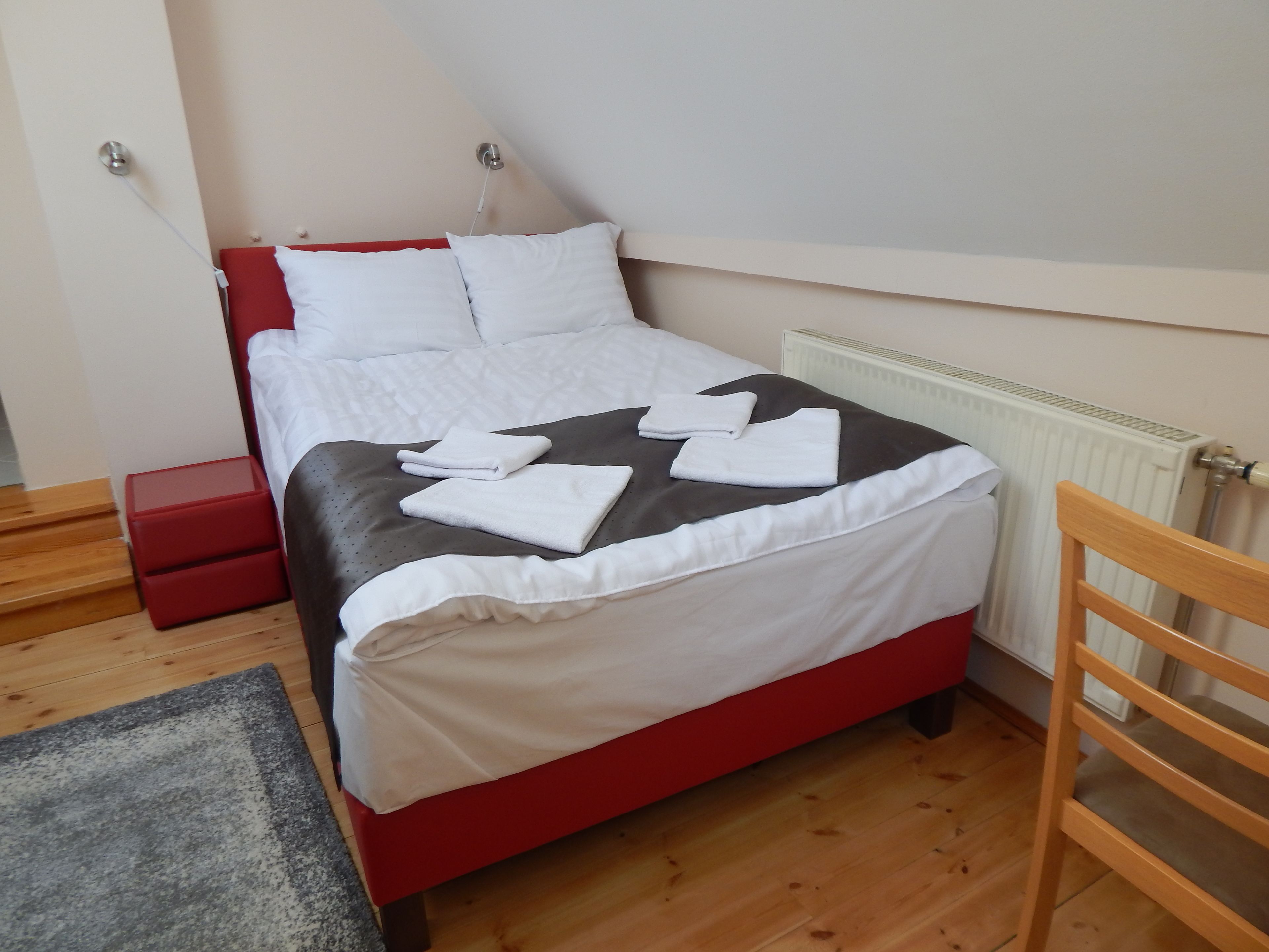 Una cama bien tendida con sábanas blancas y un detalle en negro, una mesita de noche roja y un suelo de madera.