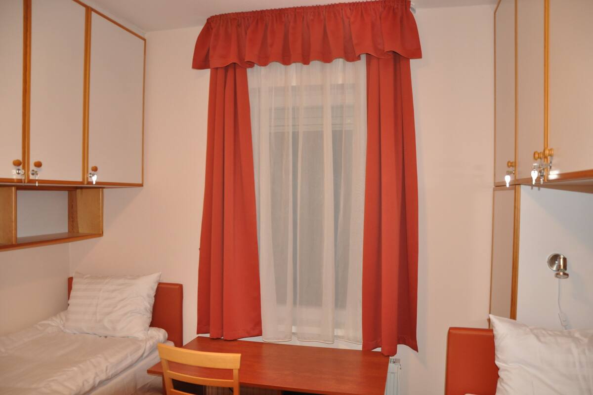 A room with two beds, a wooden table, and a window with red curtains.