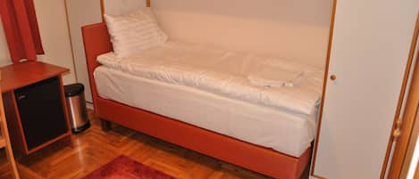 Una cama individual con estructura de madera y ropa de cama blanca, una mesita de noche pequeña, un bote de basura y una alfombra roja sobre un piso de madera.
