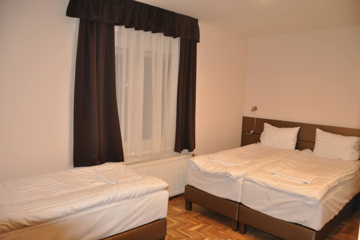 A room with two single beds, a window with curtains, a radiator, and a wooden floor.