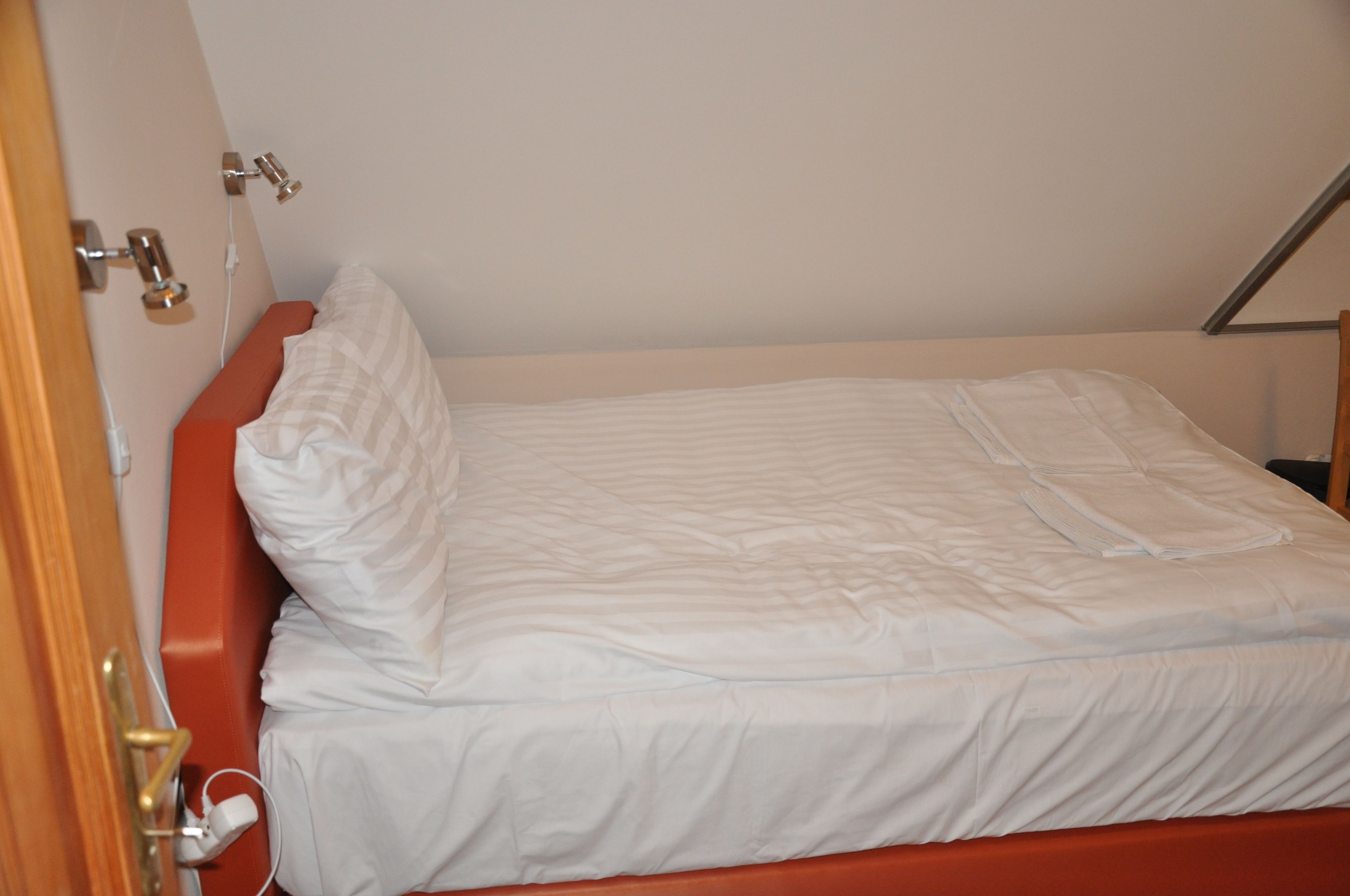 Una cama individual con colchón blanco y cabecera roja en una habitación con paredes blancas y puerta de madera.