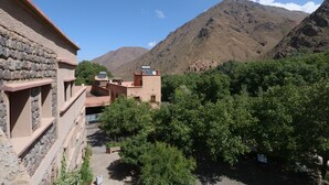 Un edificio tradicional con paredes de piedra y techo plano, rodeado de abundante vegetación y montañas al fondo.