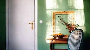 A room with a wooden floor, a green wall, a white door, a striped chair, a small table with a bowl of fruit, and a mirror reflecting a window with sunlight. - Schloss Lanke (Wandlitz)