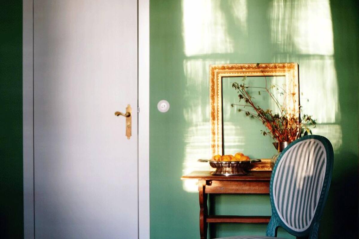 Habitación con piso de madera, pared verde, puerta blanca, una silla a rayas, una mesita con un frutero y un espejo que refleja una ventana con luz solar.