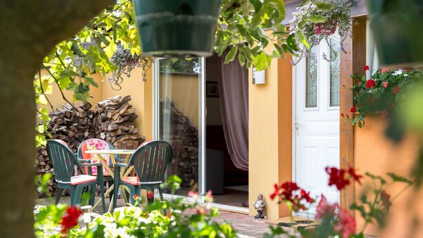 Een terras buiten met een tafel en stoelen, een stapel houtblokken, en een deur naar een binnenruimte.