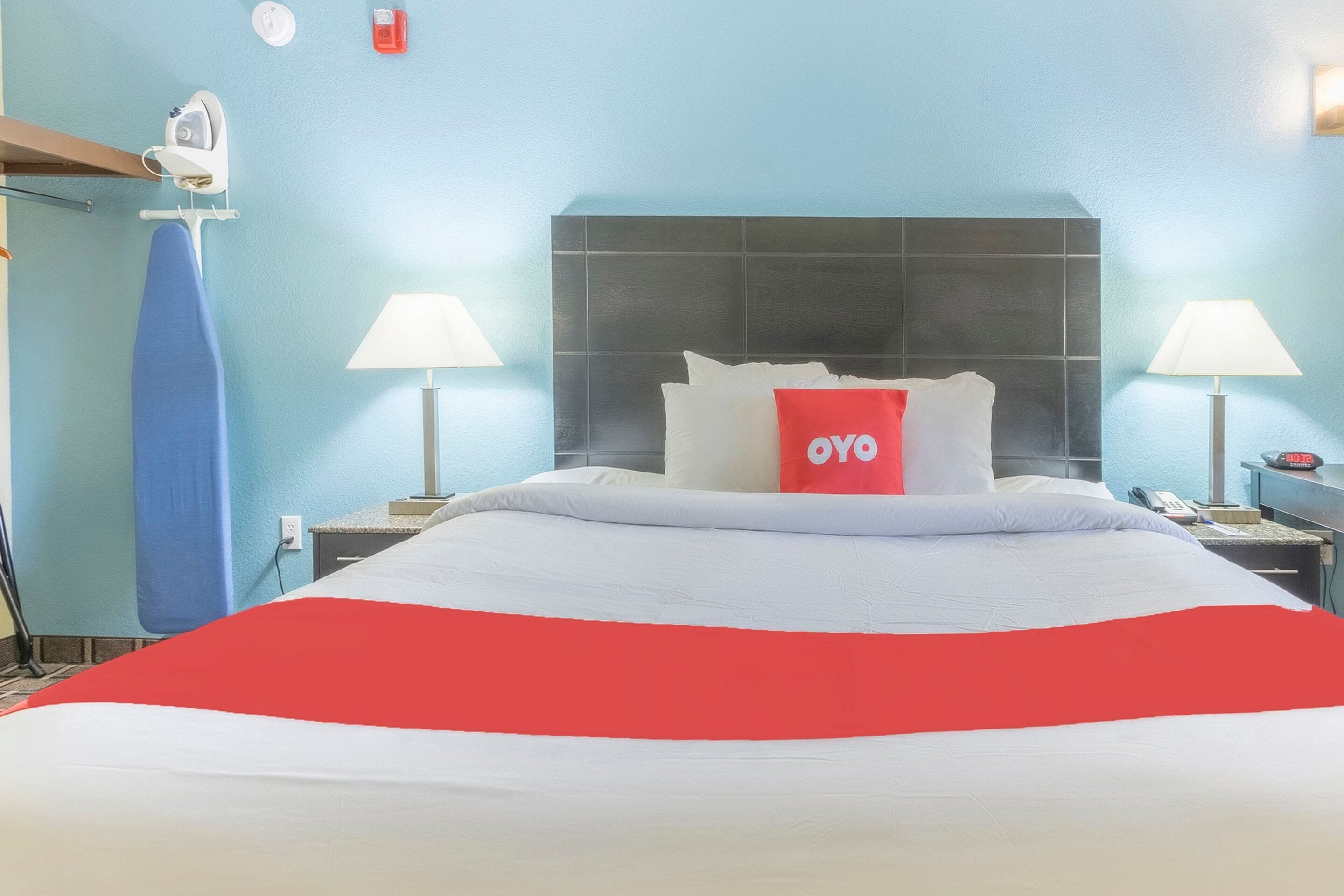 A hotel room with a bed, two bedside lamps, a red pillow, a blue ironing board, and a wall-mounted heater.