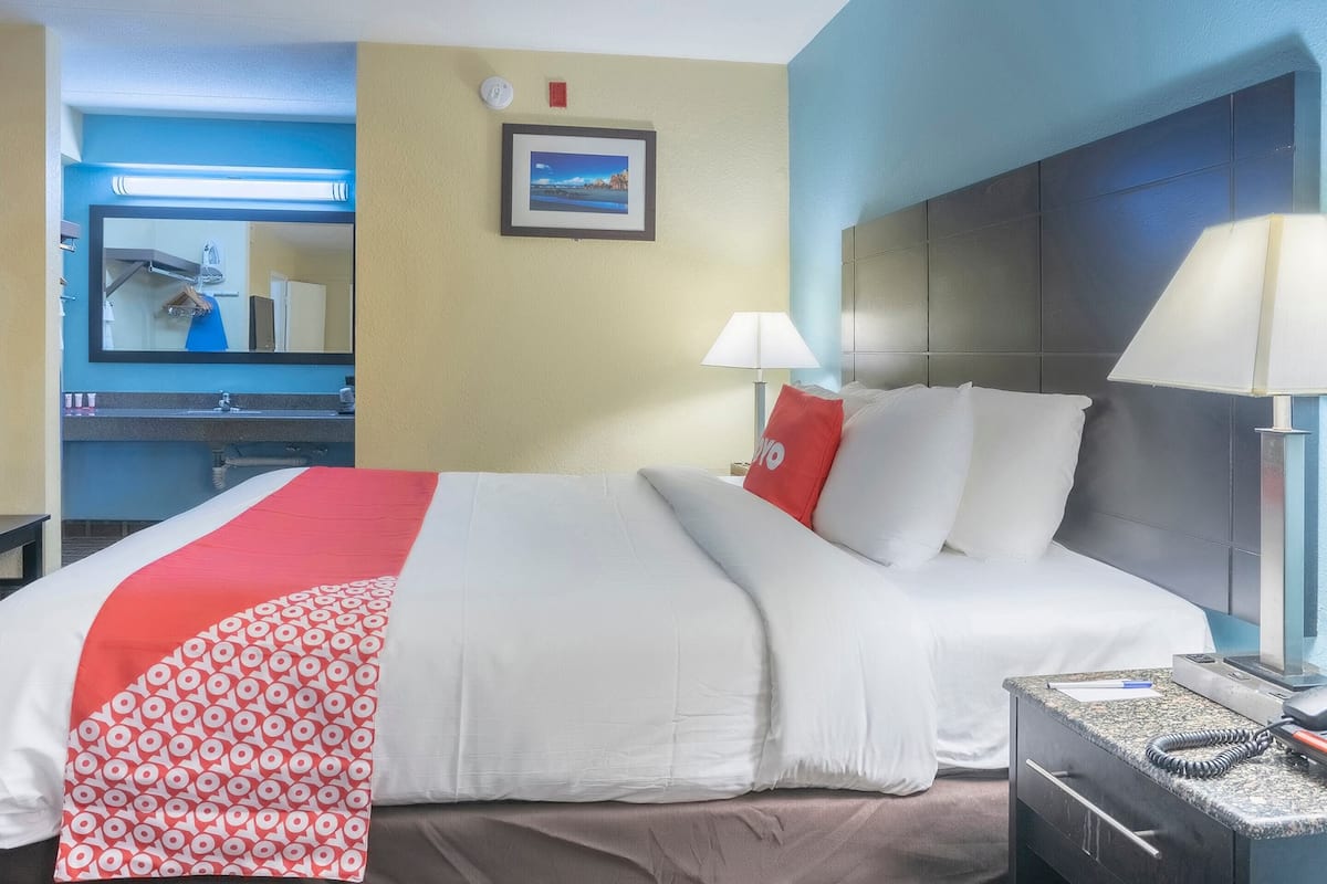 Habitación de hotel con una cama, lámparas de noche, una mesita de noche, un espejo y un cuadro en la pared.
