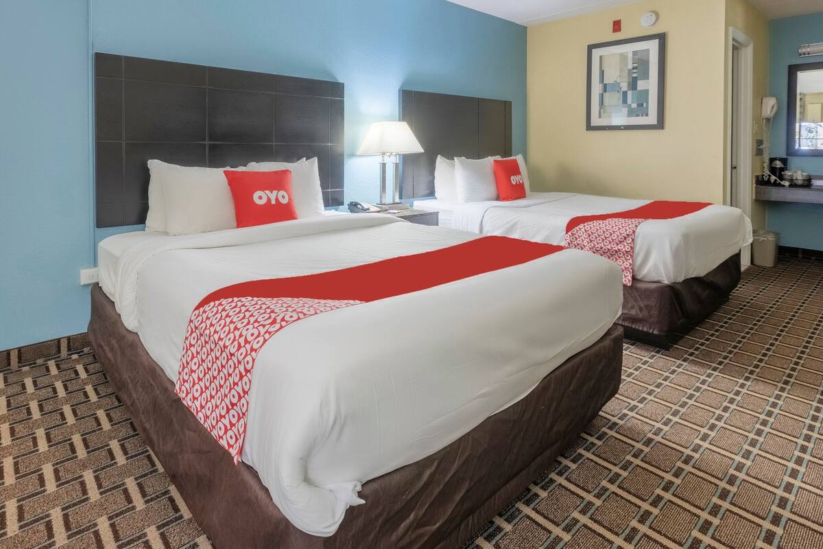 Habitación de hotel con dos camas, cada una con ropa de cama blanca y detalles rojos, una mesita de noche con lámpara, un cuadro en la pared y una alfombra estampada.