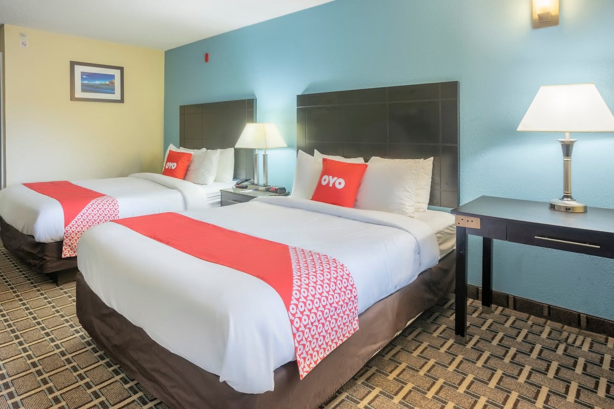 Una habitación de hotel con dos camas, cada una con colcha roja y blanca, una mesita de noche con lámpara y un escritorio con lámpara.