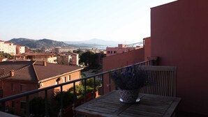 Un balcon orné d’un pot de fleurs, donnant sur un quartier résidentiel avec des bâtiments aux toits de tuiles et offrant une vue lointaine sur des collines.