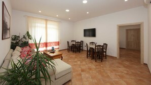 Un salon avec un canapé, une table basse et un téléviseur fixé au mur. Plusieurs chaises sont disposées autour d’une table à manger.