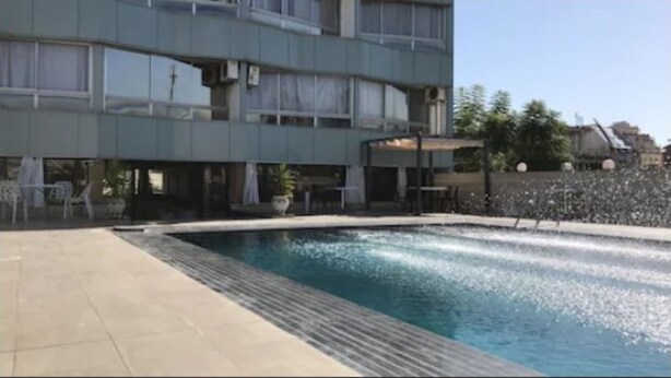 Une piscine avec une terrasse carrelée, un bâtiment doté de balcons et du mobilier d’extérieur.