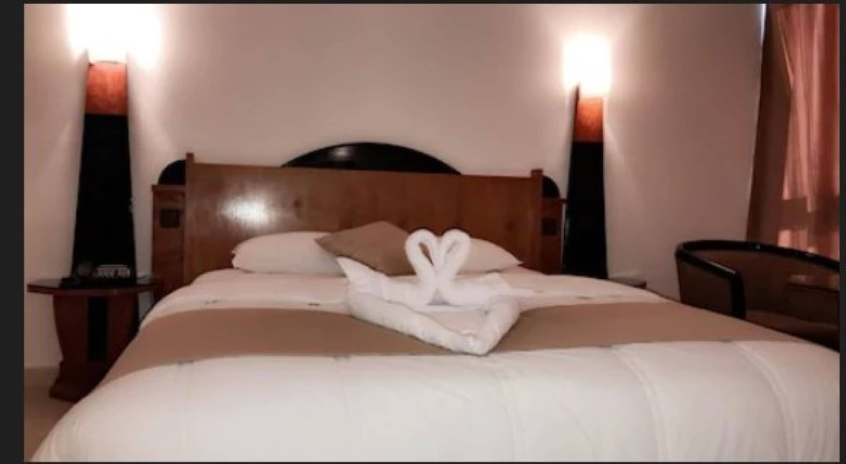 A neatly made bed with a towel arranged in a heart shape, a wooden headboard, and two bedside lamps.