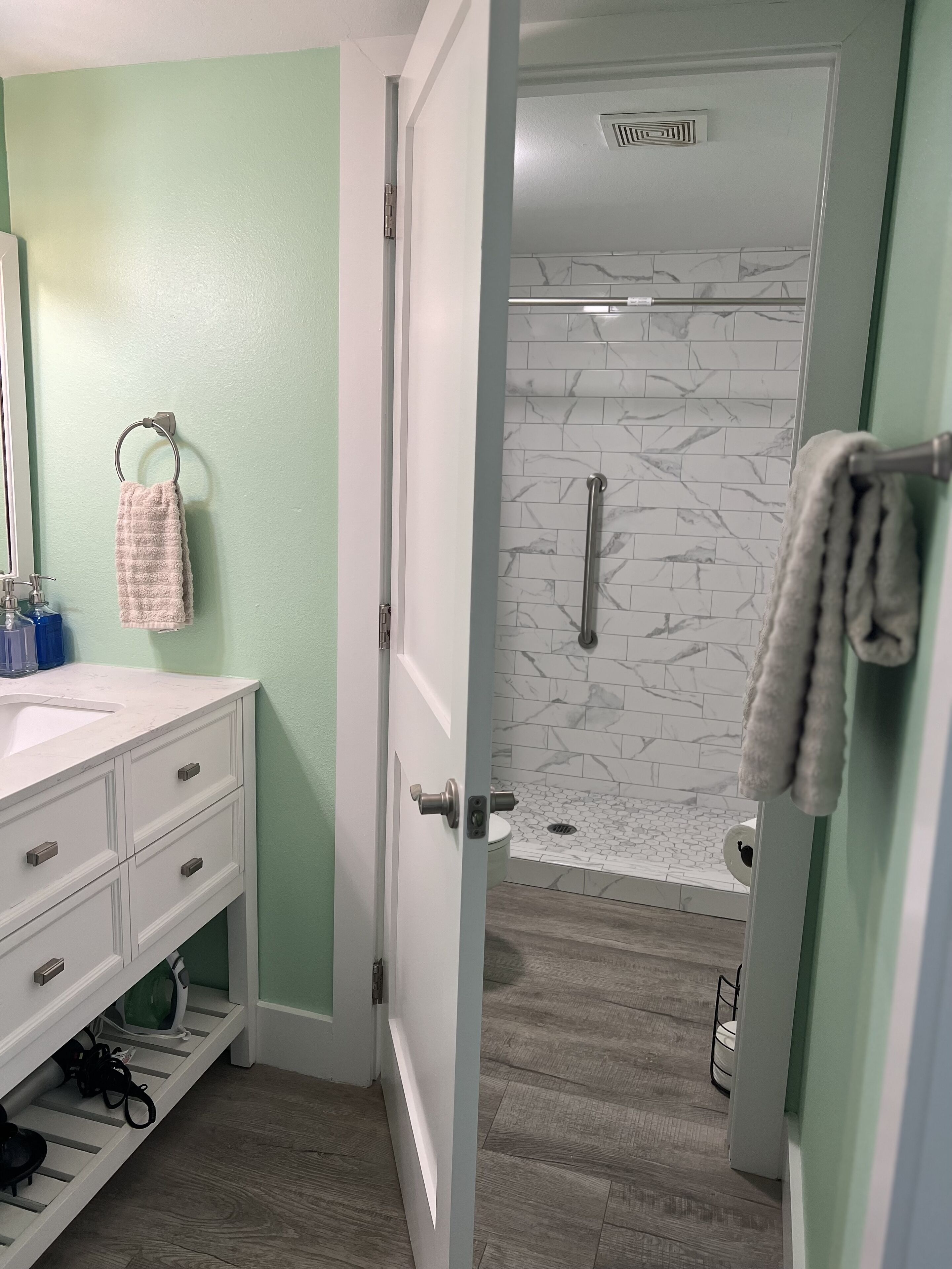 A bathroom with a white vanity, a towel rack, a shower with a marble wall, and a wooden floor.