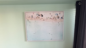 Un sofá gris con un cojín que dice "BEACH" en letras doradas, una pequeña mesita de madera y un cuadro abstracto grande en la pared.
