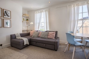 A modern living room with a grey sectional sofa, two blue chairs, a white table, and a window with white curtains.