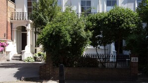Un immeuble blanc à plusieurs étages, doté d’une façade en briques, d’une clôture en métal noir et d’un arbre devant.