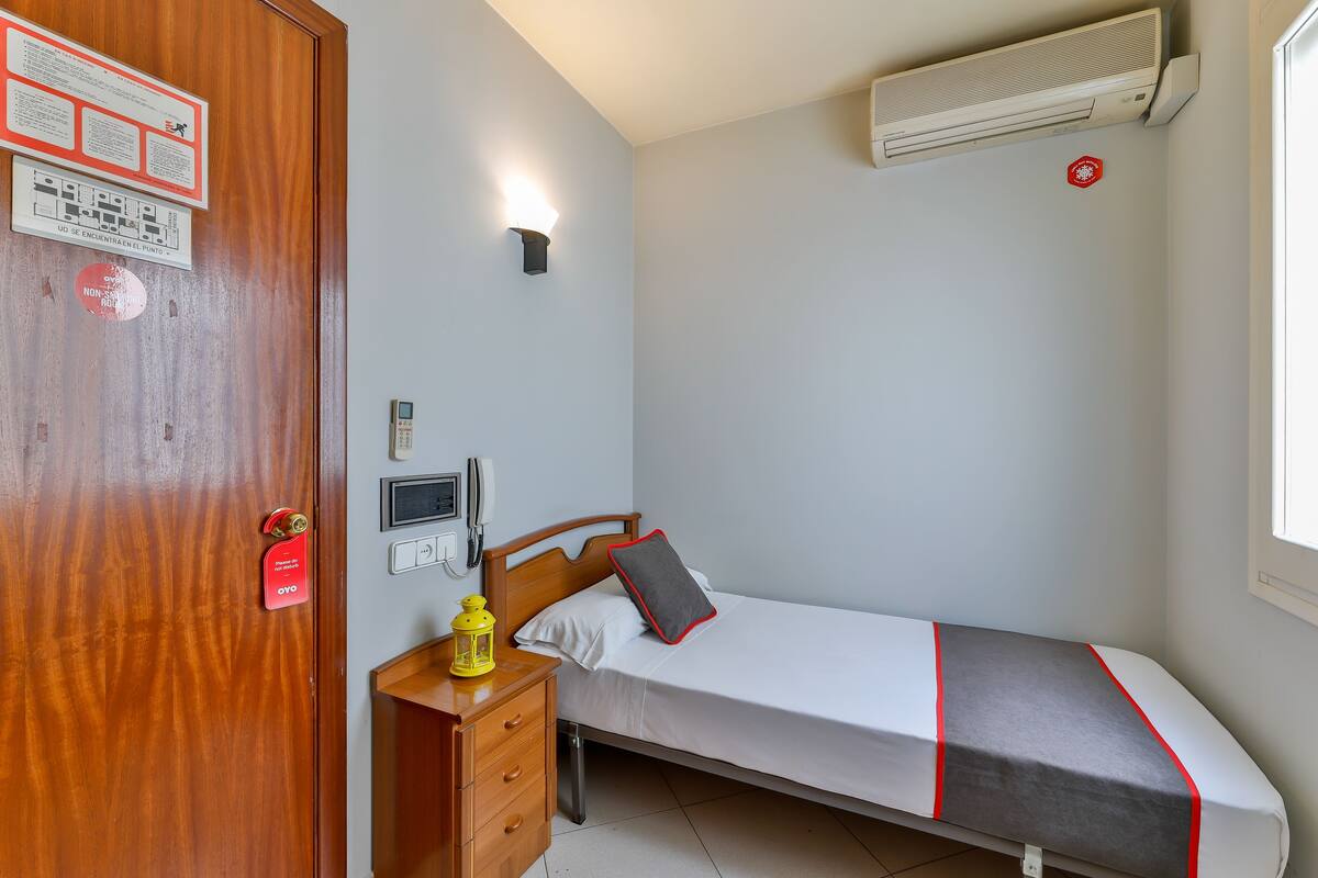 A small, neatly arranged bedroom with a single bed, a bedside table with a lamp, a wooden door, and a wall-mounted air conditioner.