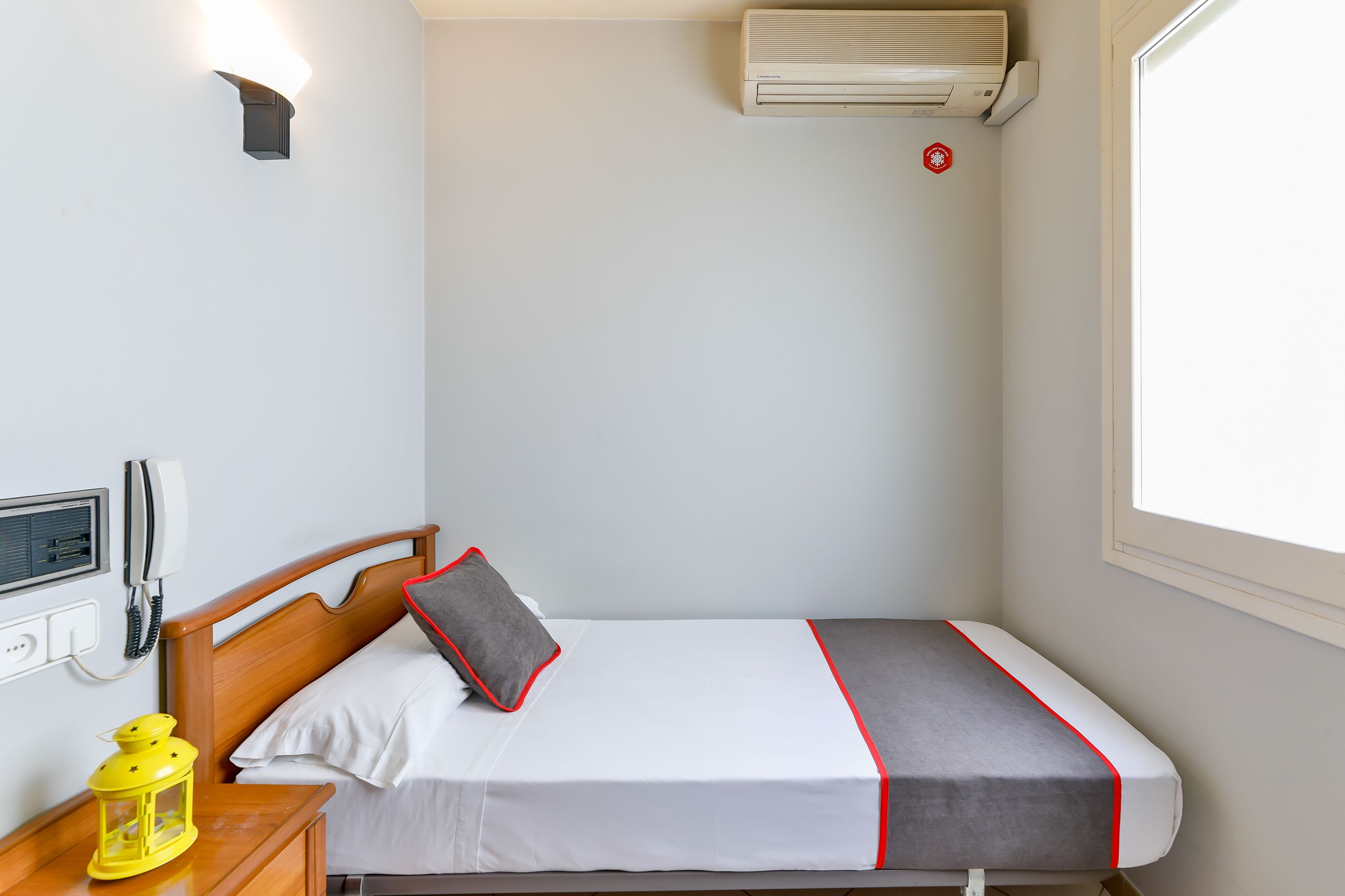 Una cama individual con cabecera de madera, una mesita de noche con un farol amarillo, un teléfono en la pared y una ventana con vistas.