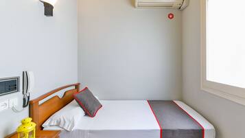 Una cama individual con cabecera de madera, una mesita de noche con un farol amarillo, un teléfono en la pared y una ventana con vistas.