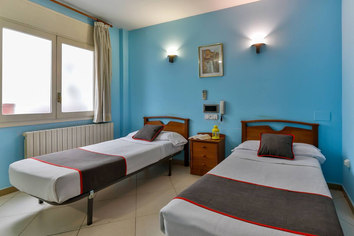 A room with two beds, a window with curtains, a bedside table with a lamp, and a wall-mounted light fixture.