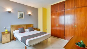 Une chambre à coucher bien rangée, avec un grand lit, une armoire en bois, des tables de chevet et une petite table sur laquelle se trouve un bol de fruits.