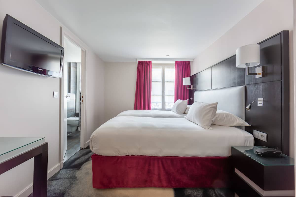 A hotel room with a bed, a TV, a bedside table, and a window with curtains.