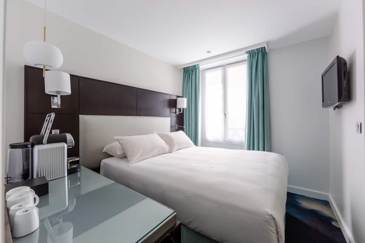 A hotel room with a bed, a desk with a lamp, a television, and a window with curtains.