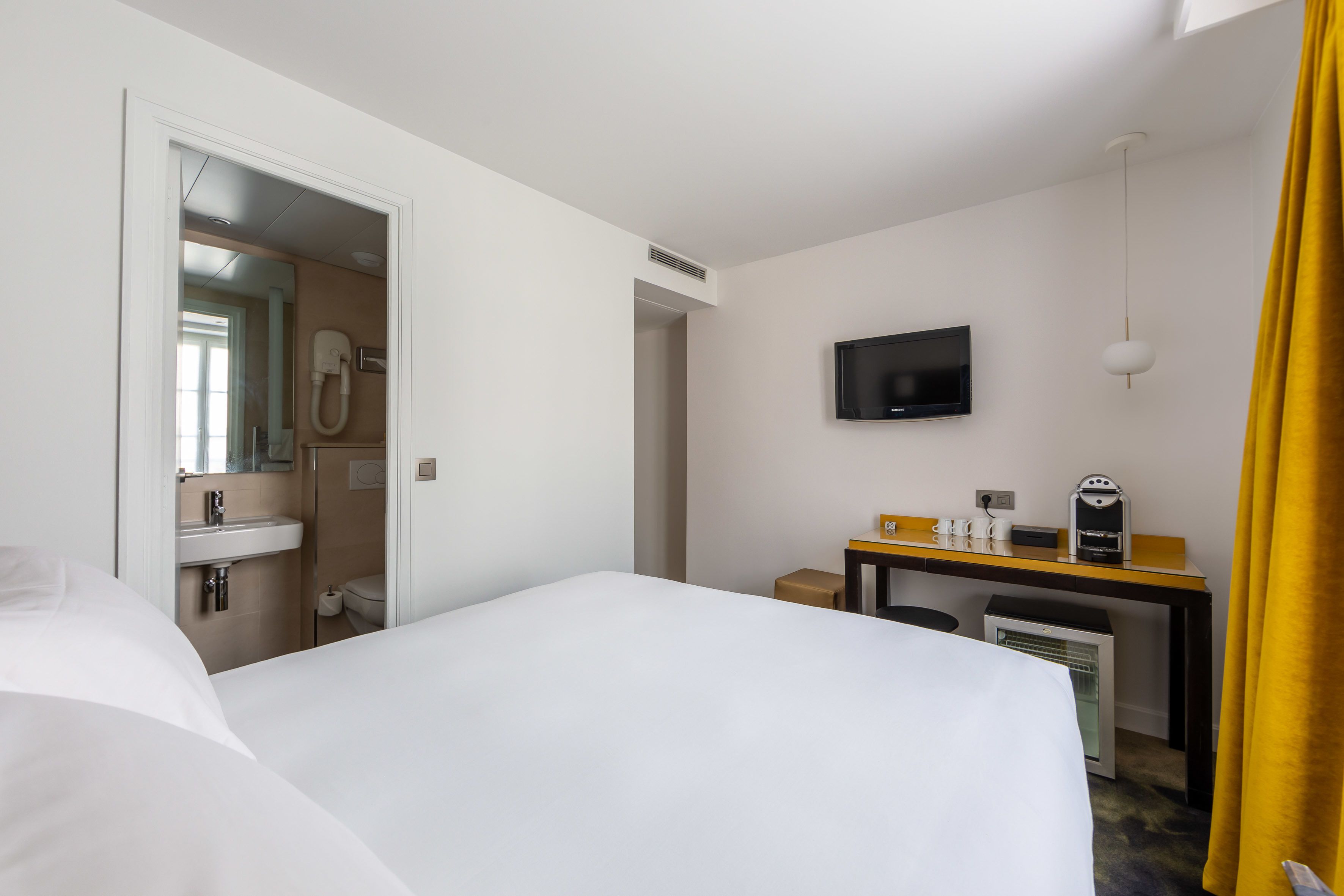 A hotel room with a bed, a desk with a computer, a TV, and a bathroom visible through an open door.