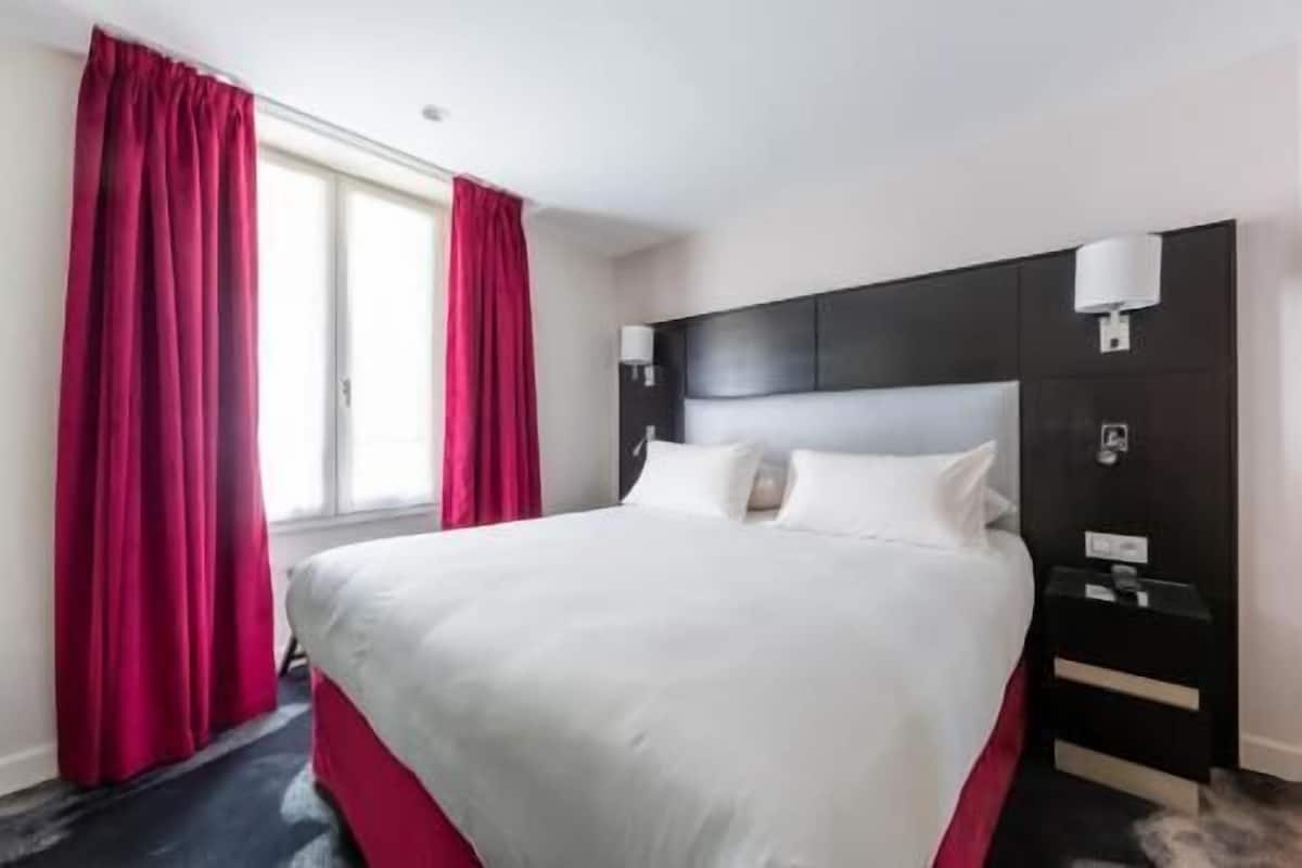 A hotel room with a large bed, a dark headboard, two bedside tables with lamps, and red curtains.