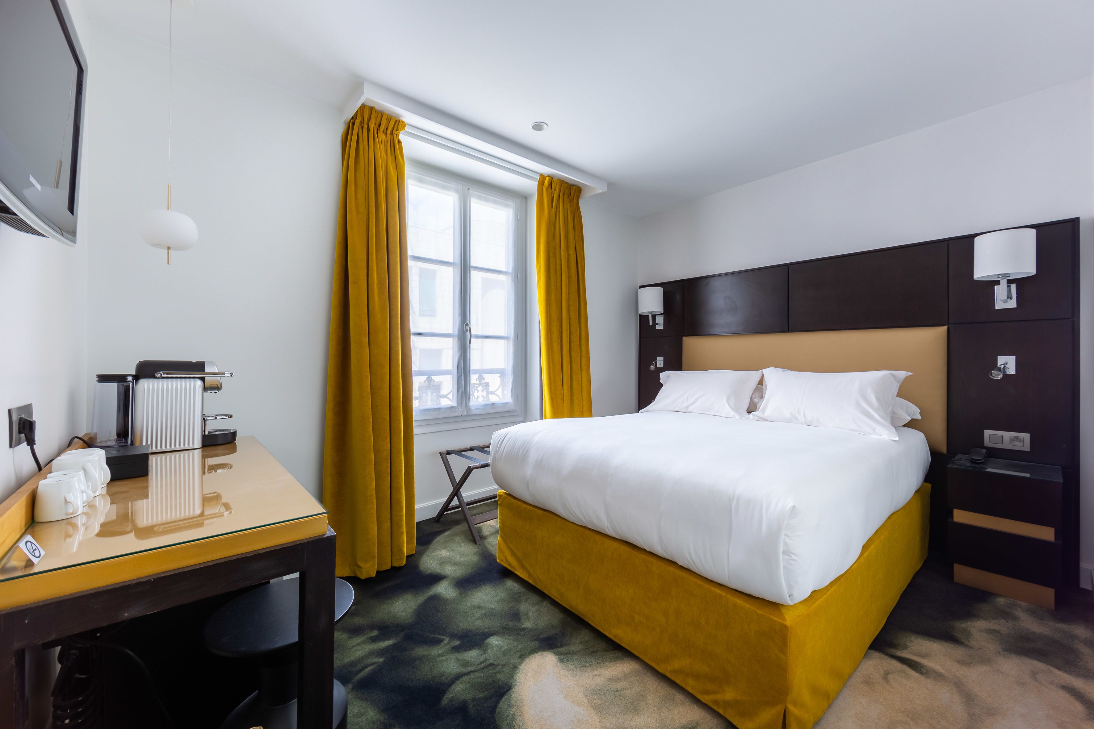 A hotel room with a bed, a desk with a computer, a lamp, and a window with curtains.