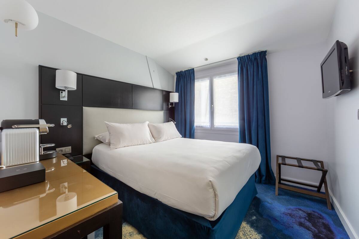 A hotel room with a large bed, bedside tables, a desk with a lamp, a TV mounted on the wall, and a window with curtains.