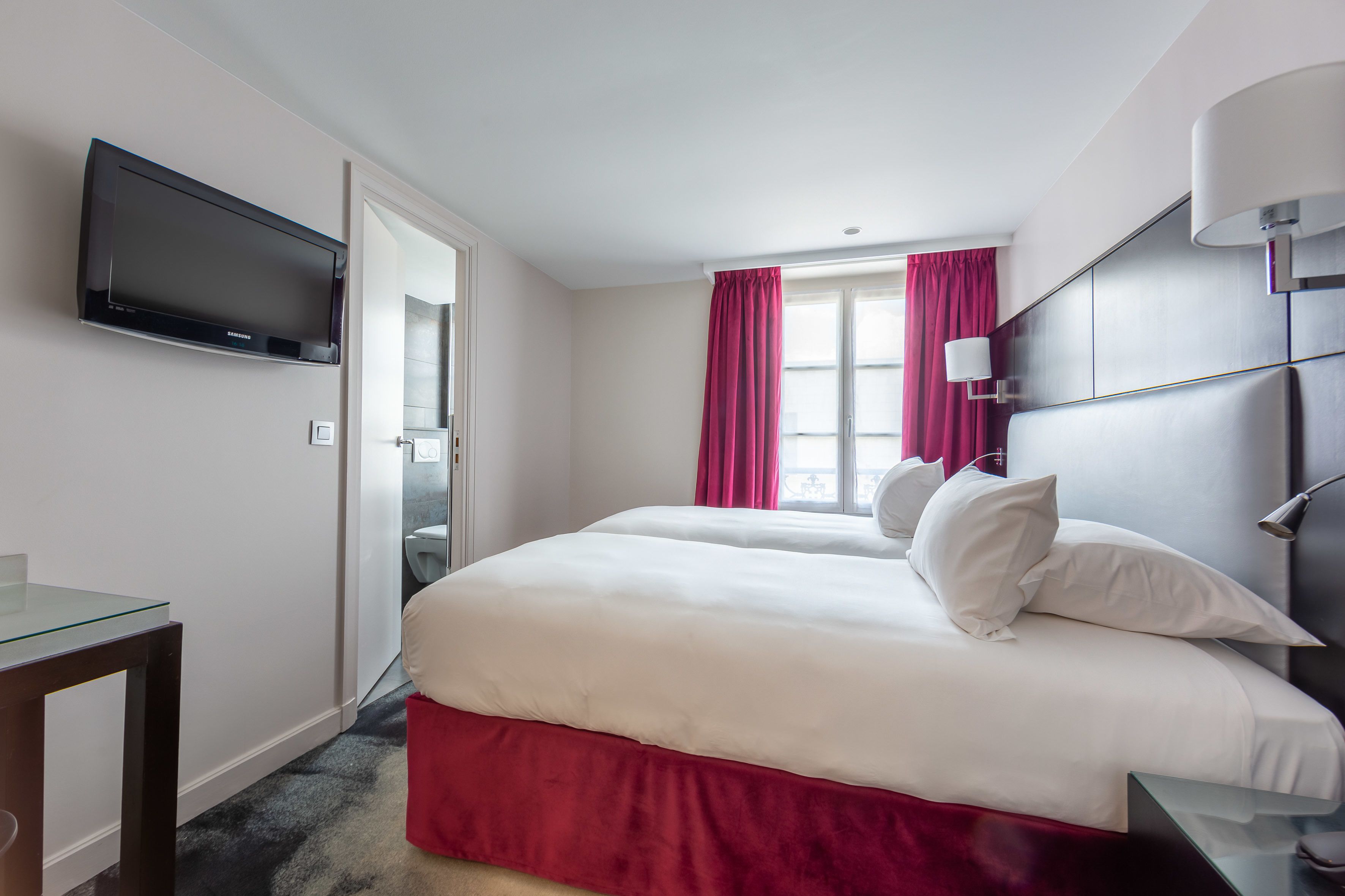 A hotel room with a bed, a TV, a desk, and a window with curtains.