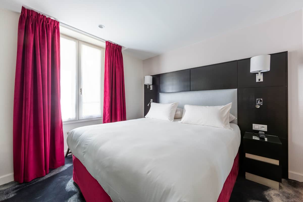 A hotel room with a large bed, red curtains, a dark headboard, bedside lamps, and a nightstand.