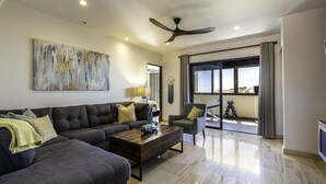 A modern living room with a sectional sofa, a coffee table, and a TV. There is a ceiling fan, a large window with curtains, and a view of the outdoors.