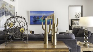 A modern living room with a wooden coffee table, a metal wine rack, a cacti sculpture, a grey sofa, and a framed painting of a coastal landscape.