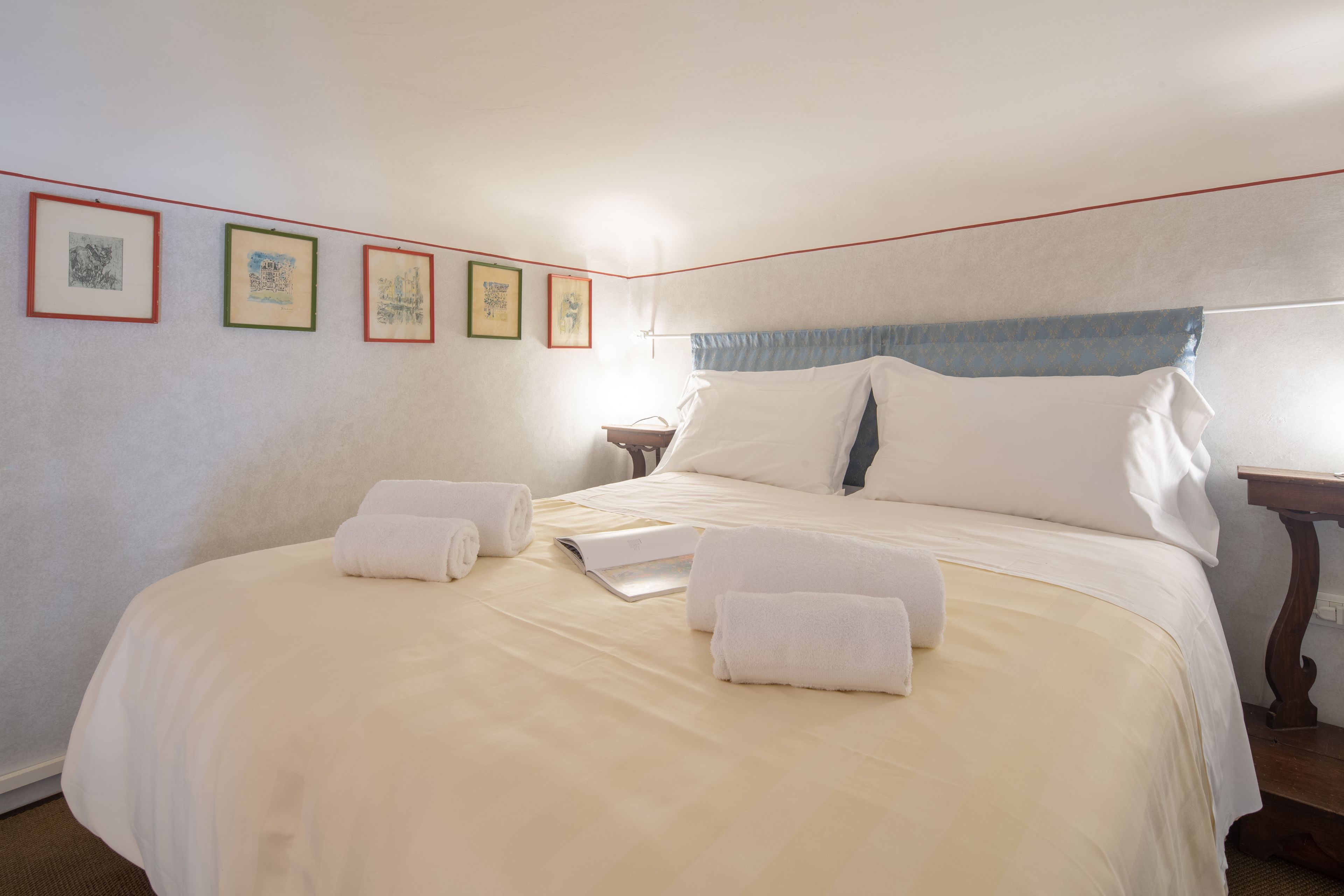 A neatly made bed with white linens and towels, a bedside table with a book, and framed pictures on the wall.