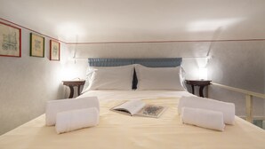 Una cama bien hecha con sábanas blancas y dos toallas, una mesita de noche con lámpara, y cuadros en la pared.
