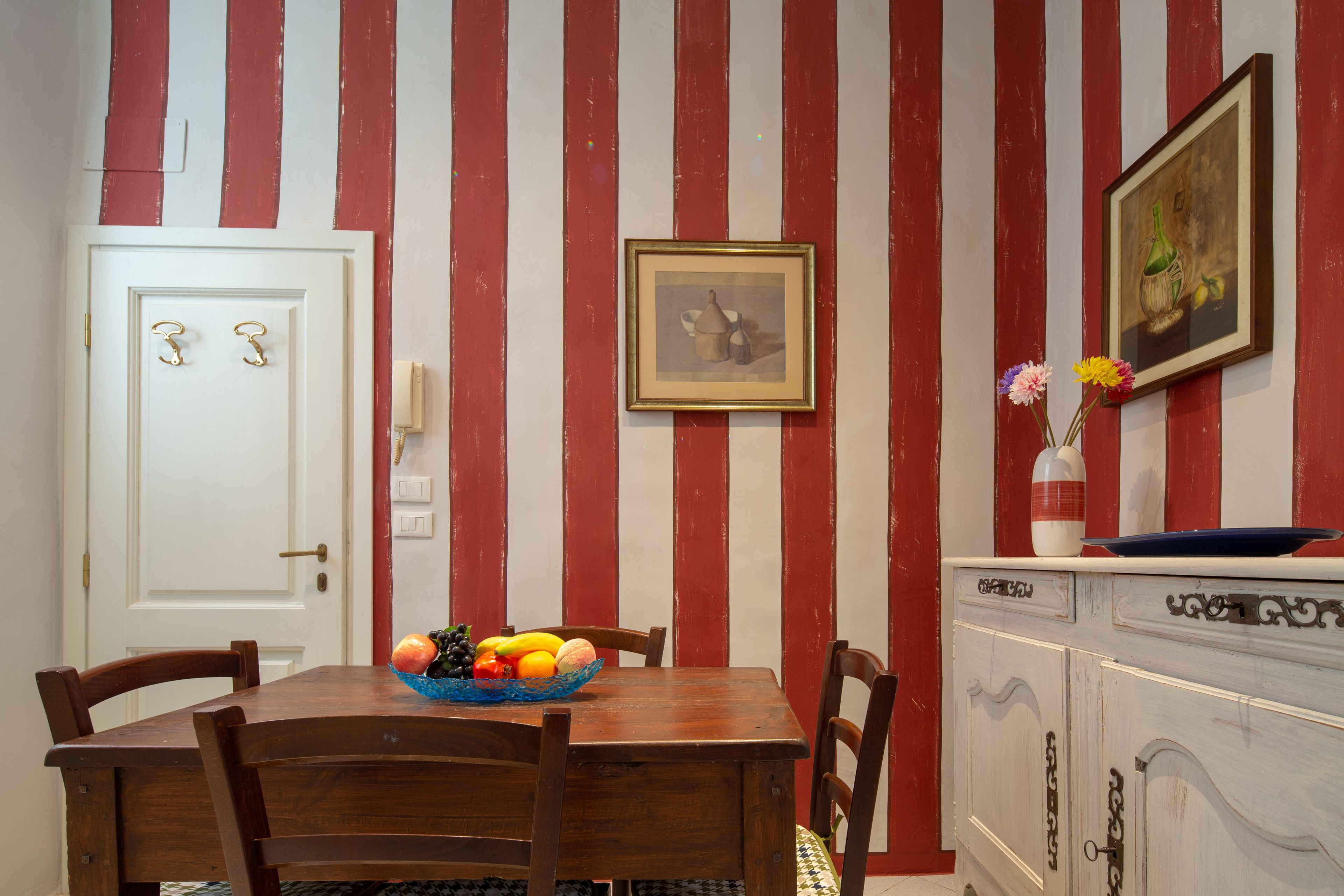 Un comedor con una mesa de madera, sillas y un frutero. Hay una puerta blanca con manija y un cuadro en la pared.