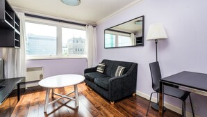 A modern living room with a dark grey sofa, a white round table, a black chair, and a floor lamp. A large window provides a view of buildings outside. - Fitzrovia Apartments by Allô Housing (London)