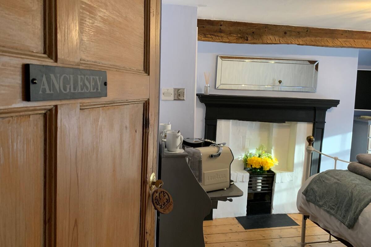 Une porte en bois sur laquelle est accrochée une plaque portant l’inscription « Anglesey », une cheminée avec un manteau de couleur blanche, et une petite table sur laquelle se trouvent une bouilloire et des fleurs.