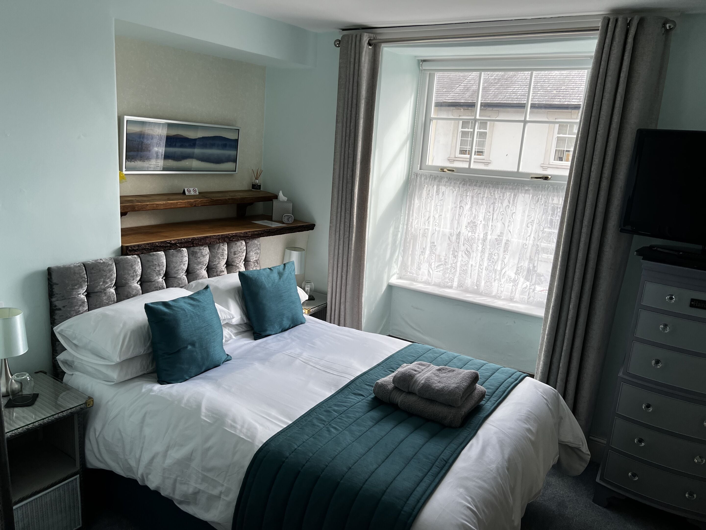 A neatly made bed with a teal bedspread and pillows, a mounted artwork above the bed, a bedside table with a lamp, and a window with curtains.