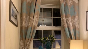 Un sillón beige con un cojín azul, una mesita de madera, una lámpara y una ventana con cortinas.