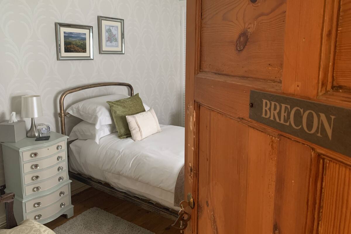 Une chambre à coucher avec un lit, une table de chevet, une lampe, deux tableaux encadrés accrochés au mur et une porte en bois sur laquelle est apposée une plaque portant l’inscription « BRECON ».