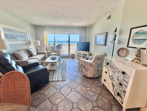 Una sala de estar con sofá, sillón, mesa de centro, televisor y vista a la playa a través de la puerta de cristal.